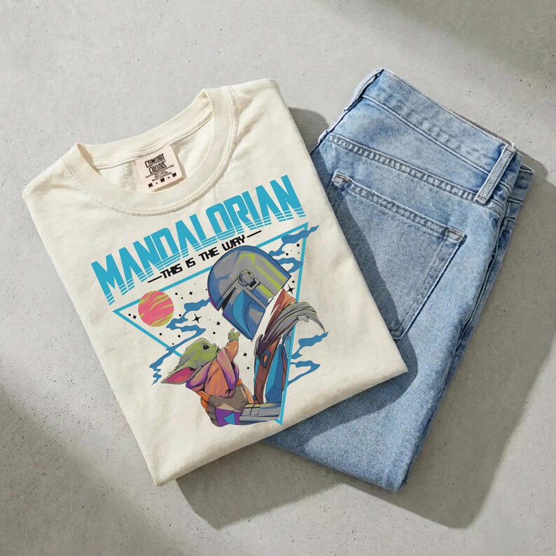Pu&ograve; includere: Una t-shirt a maniche corte color crema con una stampa grafica del Mandaloriano e Grogu. Il design include il testo "MANDALORIAN" e "THIS IS THE WAY" in blu, su un triangolo geometrico con stelle e un pianeta.