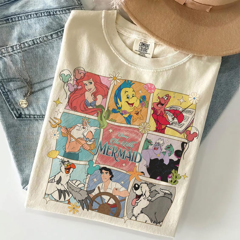 May include: Cream-coloured crew neck t-shirt featuring a colourful graphic of Disney's The Little Mermaid characters. The design includes Ariel, Flounder, Sebastian, King Triton, Prince Eric, and Ursula. The shirt is paired with light blue jeans and a necklace.