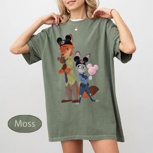 Pu&ograve; includere: T-shirt verde muschio con un'illustrazione a cartoni animati di Nick Wilde e Judy Hopps di Zootropolis, con orecchie di Topolino. La maglietta ha una vestibilit&agrave; ampia e maniche corte. La parola "Moss" &egrave; stampata in basso a sinistra.