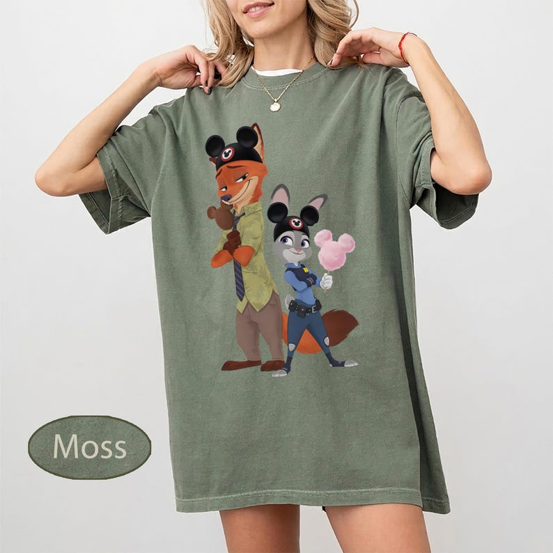 Pu&ograve; includere: T-shirt verde muschio con un'illustrazione a cartoni animati di Nick Wilde e Judy Hopps di Zootropolis, con orecchie di Topolino. La maglietta ha una vestibilit&agrave; ampia e maniche corte. La parola "Moss" &egrave; stampata in basso a sinistra.