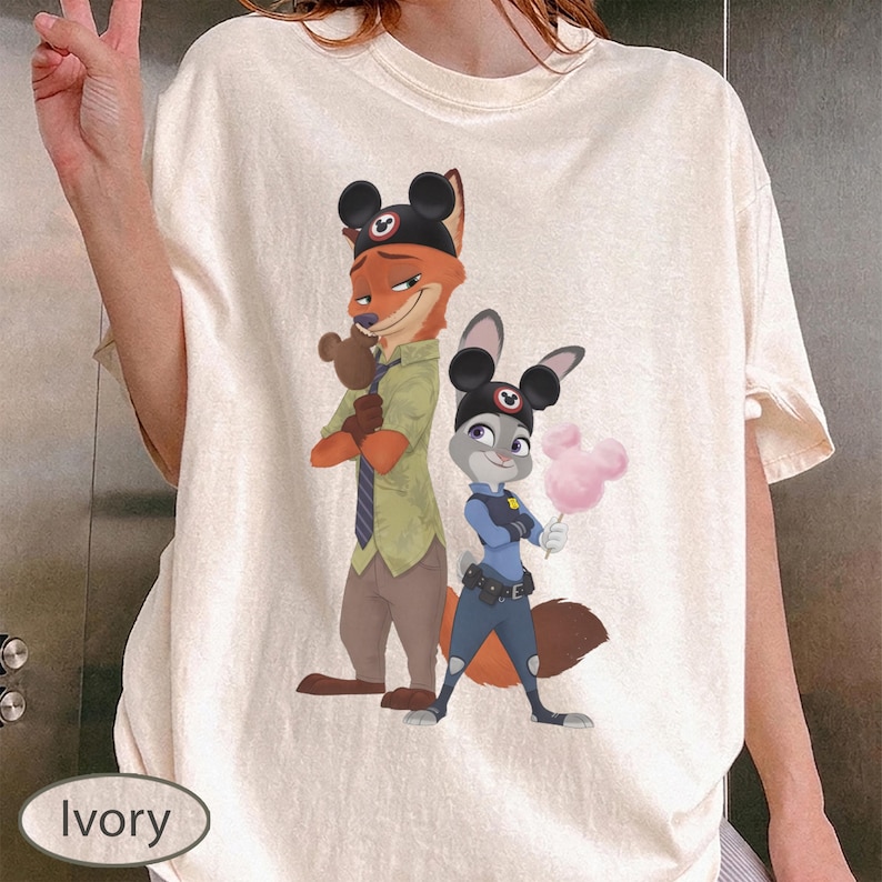 Pu&ograve; includere: T-shirt color avorio con un'illustrazione di Nick Wilde e Judy Hopps di Zootropolis, con orecchie di Topolino. Nick indossa una camicia verde e Judy un'uniforme da poliziotta. Judy tiene in mano zucchero filato rosa.