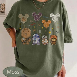 May include: Moss green t-shirt featuring Star Wars characters and Mickey Mouse-shaped balloons. The design includes Chewbacca, R2-D2, BB-8, and an Ewok. The balloons are in various colors and patterns, with lightsabers and stars.