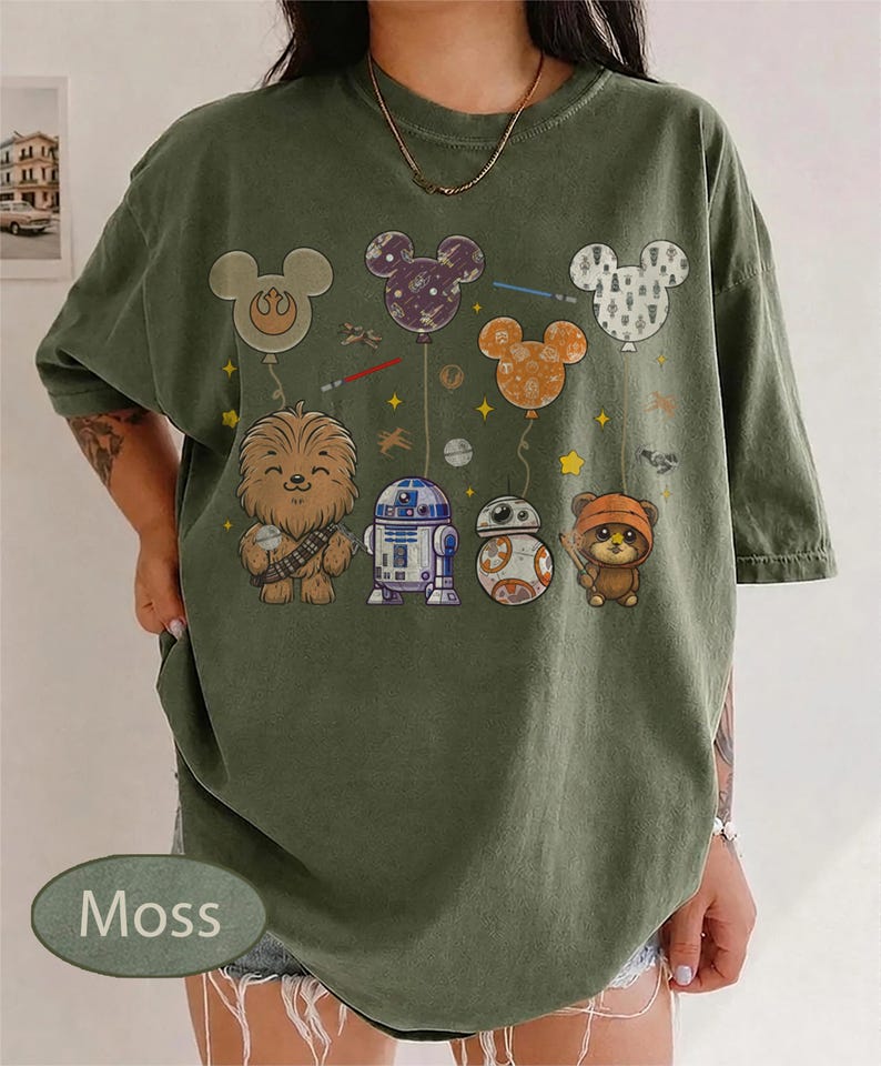 May include: Moss green t-shirt featuring Star Wars characters and Mickey Mouse-shaped balloons. The design includes Chewbacca, R2-D2, BB-8, and an Ewok. The balloons are in various colors and patterns, with lightsabers and stars.