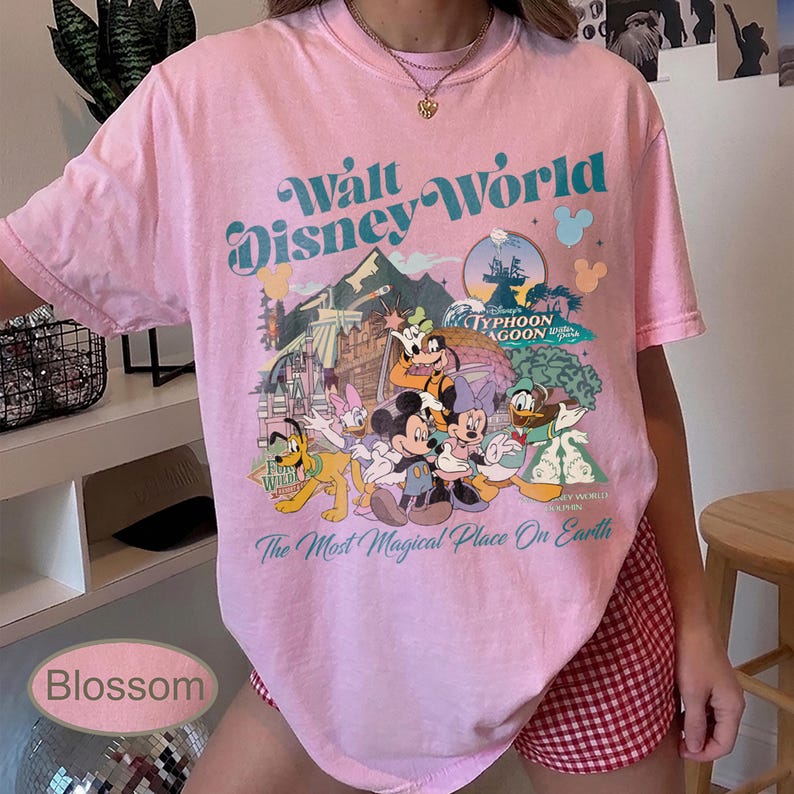 May include: Pink t-shirt with a Walt Disney World graphic. The design features Mickey Mouse, Minnie Mouse, Donald Duck, and other characters. Text includes "Walt Disney World" and "The Most Magical Place On Earth."