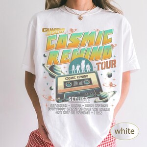 K&ouml;nnte beinhalten: Wei&szlig;es T-Shirt mit Retro-Design. Das Shirt zeigt den Text "COSMIC REWIND TOUR" und eine Kassettenband-Grafik. Das Design beinhaltet Planeten und Sterne sowie eine Setlist mit Songtiteln.