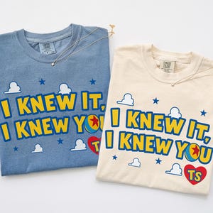 May include: A blue and a cream-colored t-shirt, each featuring the text I KNEW IT, I KNEW YOU in yellow letters with blue outlines. The design includes small white clouds, blue stars, a colorful ball, and a red heart with the letters TS inside.