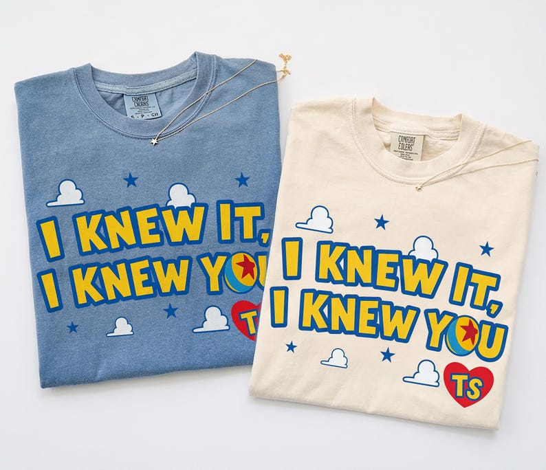 May include: A blue and a cream-colored t-shirt, each featuring the text I KNEW IT, I KNEW YOU in yellow letters with blue outlines. The design includes small white clouds, blue stars, a colorful ball, and a red heart with the letters TS inside.