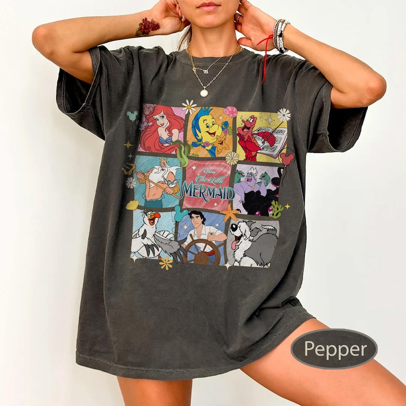 May include: Dark grey t-shirt with a colourful graphic of characters from Disney's The Little Mermaid. The design includes Ariel, Flounder, Sebastian, and other characters in a grid pattern. The shirt has a relaxed fit.
