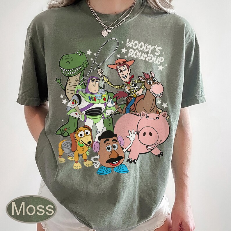 May include: Moss green t-shirt featuring a graphic of characters from the animated film "Toy Story." The design includes Woody, Buzz Lightyear, Rex, Slinky Dog, Mr. Potato Head, Hamm, and a horse. Text reads "Woody's Roundup."