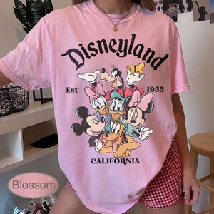 May include: Pink t-shirt featuring the word "Disneyland" in a black script, with a graphic of Disney characters. The shirt includes the text "Est 1955" and "California".