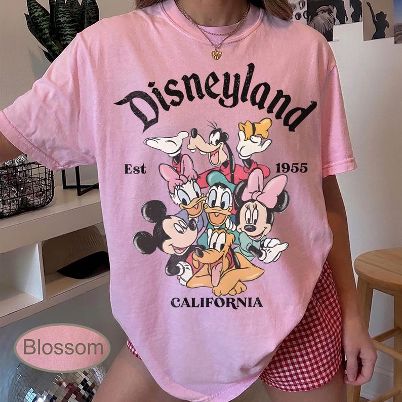May include: Pink t-shirt featuring the word "Disneyland" in a black script, with a graphic of Disney characters. The shirt includes the text "Est 1955" and "California".