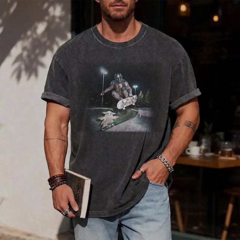 May include: A charcoal gray short-sleeve t-shirt with a graphic print featuring a character in metallic armor performing a skateboard trick in the air above a small creature on a skateboard, set against a dark outdoor night scene with streetlights.