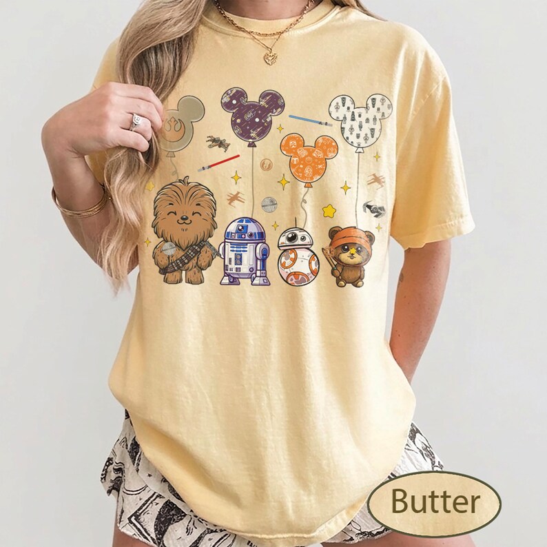 May include: Butter-colored t-shirt featuring cartoon characters from Star Wars and Mickey Mouse-shaped balloons. The design includes Chewbacca, R2-D2, BB-8, and an Ewok. The balloons are purple, orange, and white.