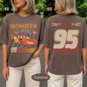 May include: Espresso-colored t-shirt featuring a graphic of a red race car with the number 95 and the name "McQueen". The front design includes checkered flags and a trophy. The back displays the number 95 in large, off-white letters.