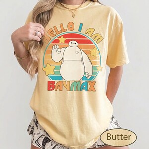 May include: A light yellow t-shirt featuring a cartoon image of Baymax from Big Hero 6. The shirt has the text "HELLO I AM BAYMAX" in a colorful design. The shirt is a casual style.