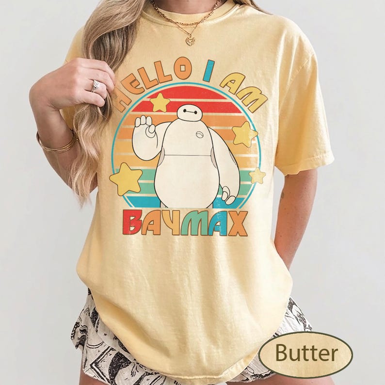 May include: A light yellow t-shirt featuring a cartoon image of Baymax from Big Hero 6. The shirt has the text "HELLO I AM BAYMAX" in a colorful design. The shirt is a casual style.