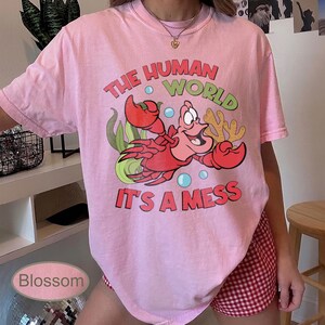 May include: Pink t-shirt featuring a cartoon crab with the text "THE HUMAN WORLD IT'S A MESS" in red, green, and blue. The graphic includes bubbles and seaweed. The shirt is a light pink color.