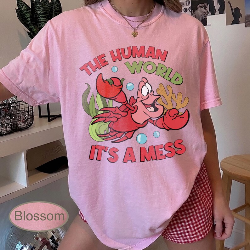 May include: Pink t-shirt featuring a cartoon crab with the text "THE HUMAN WORLD IT'S A MESS" in red, green, and blue. The graphic includes bubbles and seaweed. The shirt is a light pink color.
