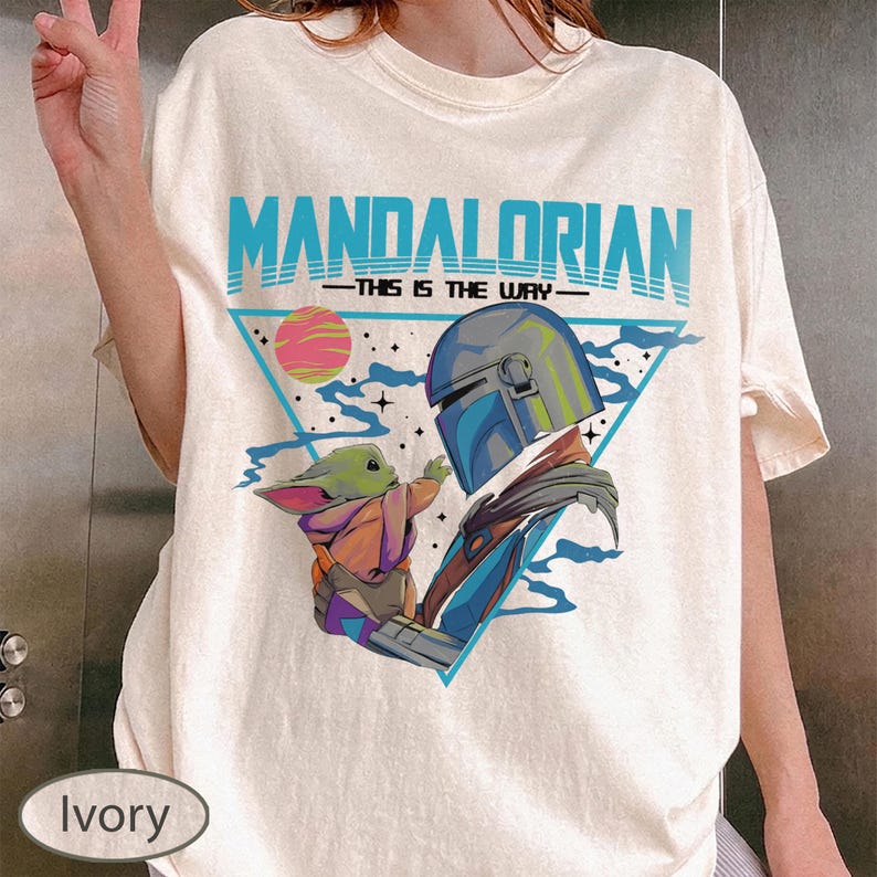 Pu&ograve; includere: Una t-shirt color avorio con una stampa grafica del Mandaloriano e Grogu. Il design include il testo "MANDALORIAN" e "THIS IS THE WAY" in blu, un pianeta rosa e un triangolo blu. L'illustrazione raffigura i personaggi in modo stilizzato.