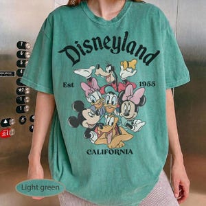 May include: Light green t-shirt featuring the word "Disneyland" in a distressed font, along with the text "Est 1955" and "CALIFORNIA". The shirt showcases a colorful graphic of Disney characters, including Mickey Mouse, Minnie Mouse, and Goofy.