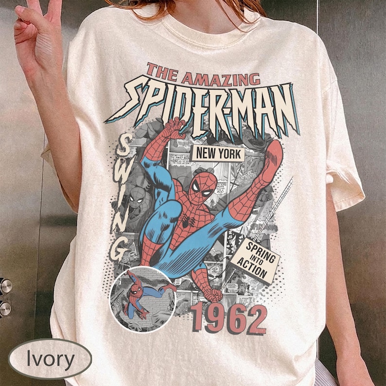 May include: Ivory-colored t-shirt featuring a vintage comic book graphic of Spider-Man. The design includes the text "THE AMAZING SPIDERMAN NEW YORK 1962" and a smaller image of Spider-Man. The shirt has short sleeves.