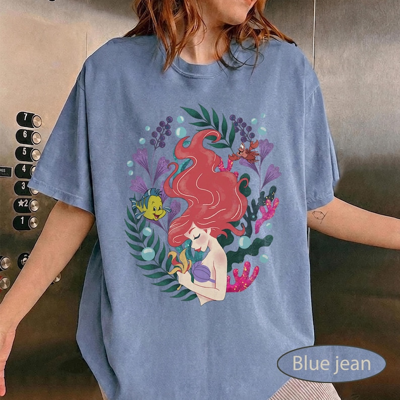 Peut inclure: T-shirt bleu jean avec un motif color&eacute; d'une sir&egrave;ne aux cheveux roux, entour&eacute;e de plantes aquatiques et de cr&eacute;atures marines. Le motif comprend un poisson jaune, un crabe et d'autres animaux marins. Le t-shirt a des manches courtes et un col rond.