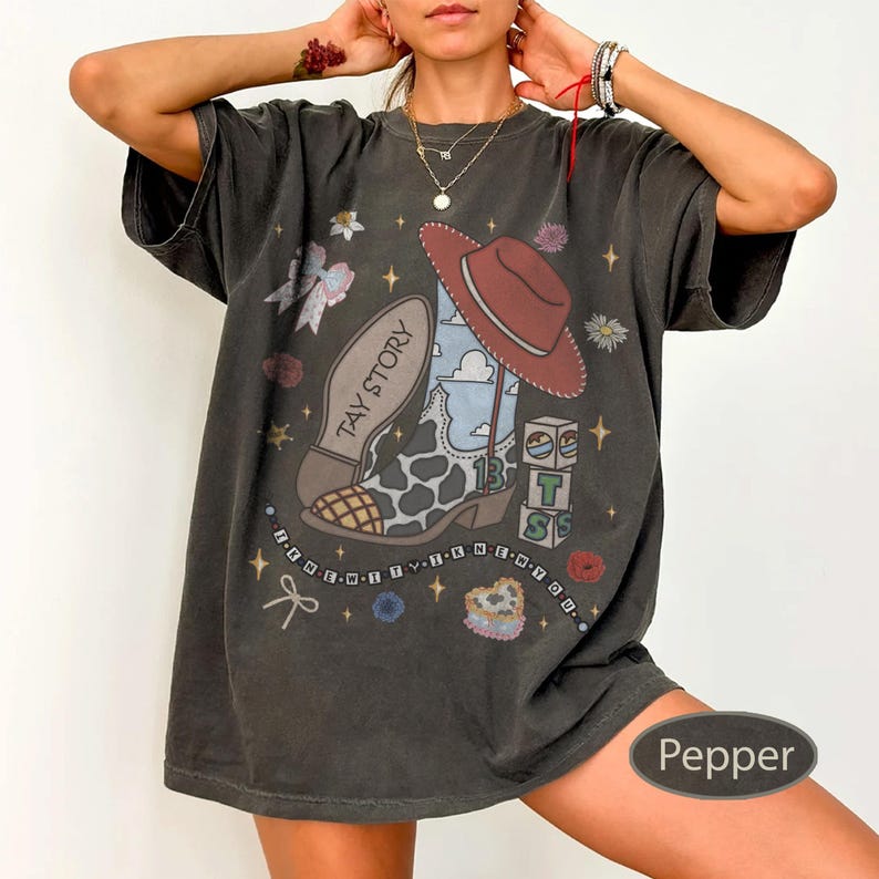 May include: A charcoal gray t-shirt featuring a graphic print of a cow-print cowboy boot, a red cowboy hat, and blocks with the text TAY STORY. The design includes stars, bows, and a beaded string graphic. The shirt has a loose, relaxed fit.