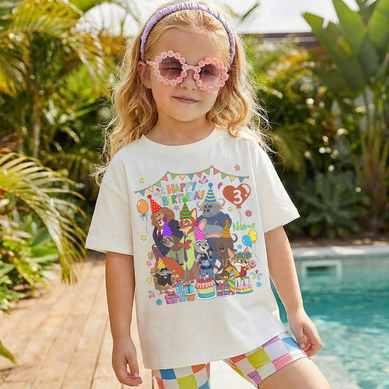May include: A young girl wearing a white t-shirt with a colorful graphic of cartoon animals and the text "HAPPY BIRTHDAY 3". She is also wearing pink flower-shaped sunglasses, a purple headband, and checkered shorts.