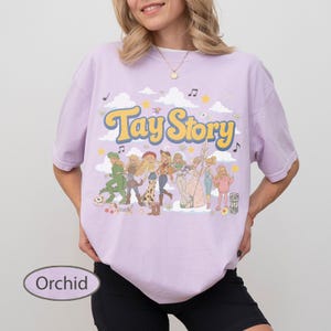 K&ouml;nnte beinhalten: Ein orchideefarbenes T-Shirt mit dem gelben Schriftzug Tay Story in einer Retro-Schriftart. Die Grafik zeigt eine Gruppe illustrierter Figuren in verschiedenen Outfits, umgeben von wei&szlig;en Wolken, gelben Sternen und schwarzen Noten auf hellem Grund.