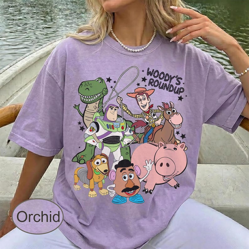 May include: A lilac-colored t-shirt featuring a graphic of characters from the animated film "Toy Story." The design includes Woody, Buzz Lightyear, Rex, Hamm, Mr. Potato Head, Slinky Dog, and Bullseye, with the text "Woody's Roundup."