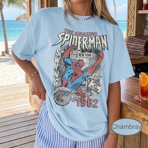 May include: Light blue t-shirt featuring a vintage comic book graphic of Spider-Man in action. The graphic includes the text "The Amazing Spider-Man," "New York," and "1962." The shirt has short sleeves and a relaxed fit.