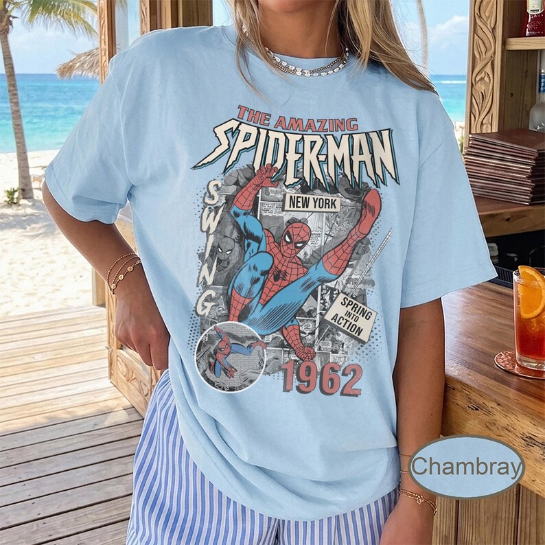 May include: Light blue t-shirt featuring a vintage comic book graphic of Spider-Man in action. The graphic includes the text "The Amazing Spider-Man," "New York," and "1962." The shirt has short sleeves and a relaxed fit.