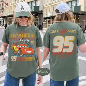 May include: Moss green t-shirt featuring a graphic of Lightning McQueen from the Cars movie. The front design includes the number 95, checkered flags, and a trophy. The back of the shirt displays the number 95 and the text "Lightning McQueen".