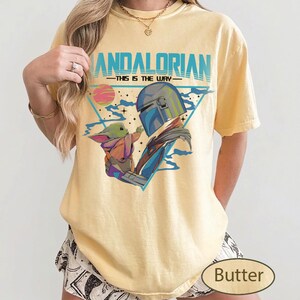 Pu&ograve; includere: Una t-shirt a maniche corte giallo pallido con una stampa grafica del Mandaloriano e di Grogu. Il testo recita MANDALORIAN THIS IS THE WAY. Il design include un triangolo blu, un pianeta rosa e stelle su uno sfondo in tessuto giallo chiaro.