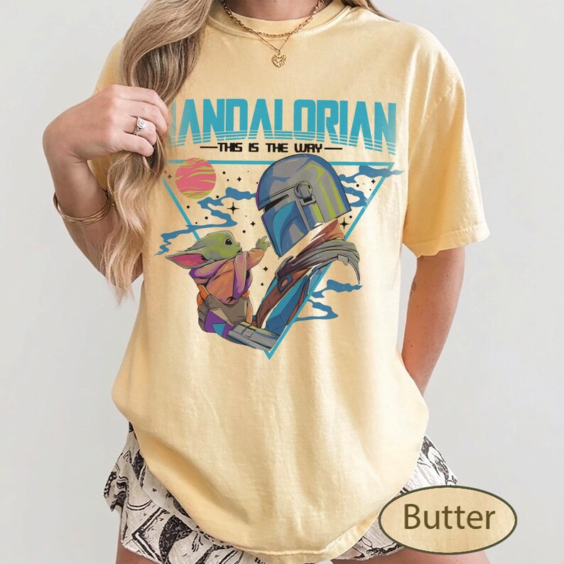Pu&ograve; includere: Una t-shirt a maniche corte giallo pallido con una stampa grafica del Mandaloriano e di Grogu. Il testo recita MANDALORIAN THIS IS THE WAY. Il design include un triangolo blu, un pianeta rosa e stelle su uno sfondo in tessuto giallo chiaro.