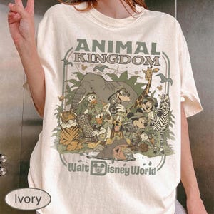 May include: Ivory-colored t-shirt featuring a graphic of Disney characters in an Animal Kingdom theme. The design includes an elephant, giraffe, tiger, zebra, and other characters. Text on the shirt reads "ANIMAL KINGDOM" and "Walt Disney World".