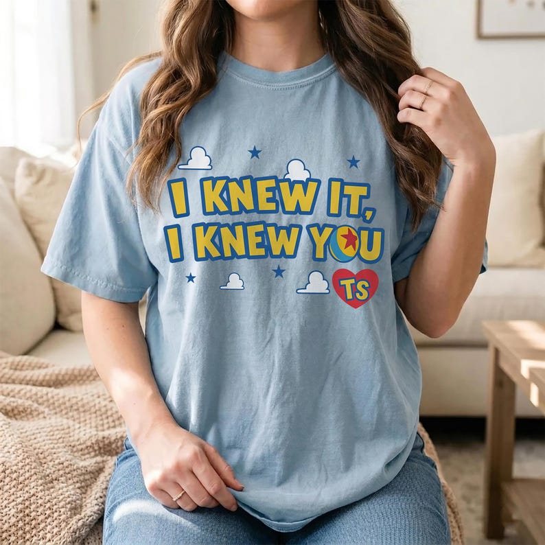 May include: A light blue short-sleeve t-shirt featuring the yellow text I KNEW IT, I KNEW YOU with a red heart containing the letters TS, a colorful ball graphic, and white cloud and blue star accents printed on the front of the garment.