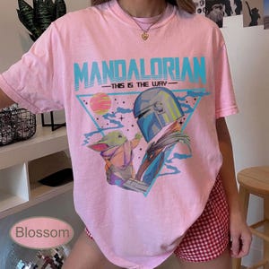 Pu&ograve; includere: Una t-shirt rosa chiaro con una stampa grafica di The Mandalorian e Grogu. Il testo recita MANDALORIAN e THIS IS THE WAY. Il design include un triangolo blu, un pianeta e stelle. La t-shirt ha un girocollo e maniche corte.