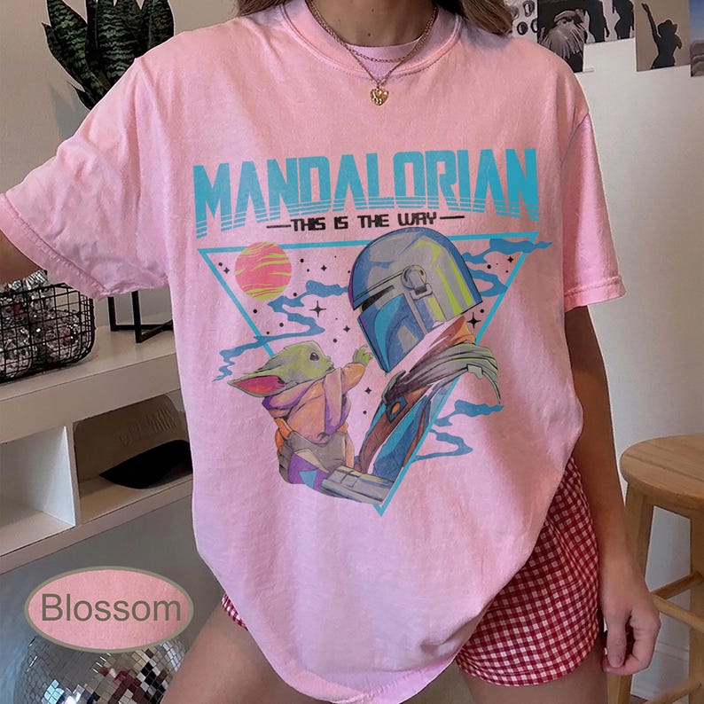 Pu&ograve; includere: Una t-shirt rosa chiaro con una stampa grafica di The Mandalorian e Grogu. Il testo recita MANDALORIAN e THIS IS THE WAY. Il design include un triangolo blu, un pianeta e stelle. La t-shirt ha un girocollo e maniche corte.