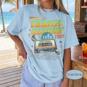 K&ouml;nnte beinhalten: Hellblaues T-Shirt mit einem Grafikdesign, das die "Cosmic Rewind Tour" bewirbt. Das Design zeigt eine Kassette, Planeten und den Text "Cosmic Rewind Tour" in Orange und Gr&uuml;n. Die Setlist enth&auml;lt Songs wie "September" und "Disco Inferno".