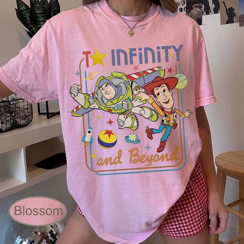 May include: Pink t-shirt featuring Buzz Lightyear and Woody from Toy Story with the words "To Infinity and Beyond." The graphic includes a star, rocket, and other colorful elements. The shirt is a light pink color.