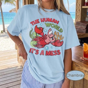 May include: Light blue t-shirt featuring a red cartoon crab with the text "THE HUMAN WORLD" and "IT'S A MESS" in a playful font. The design includes bubbles, seaweed, and coral. The shirt is a casual wear item.