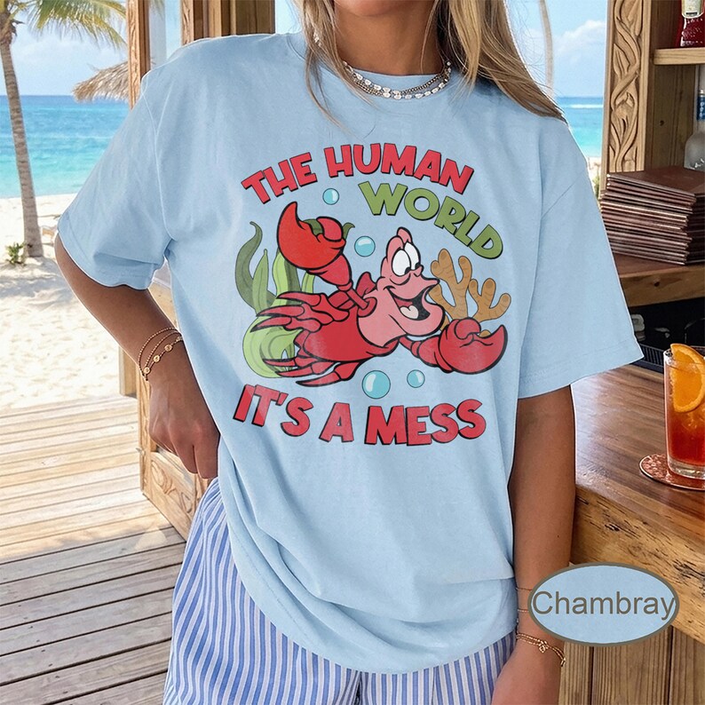 May include: Light blue t-shirt featuring a red cartoon crab with the text "THE HUMAN WORLD" and "IT'S A MESS" in a playful font. The design includes bubbles, seaweed, and coral. The shirt is a casual wear item.