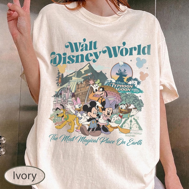 May include: Ivory-colored t-shirt featuring a colorful graphic of Disney characters and the text "Walt Disney World." The design includes Mickey Mouse, Minnie Mouse, Donald Duck, and Goofy. The shirt also has the text "The Most Magical Place On Earth."