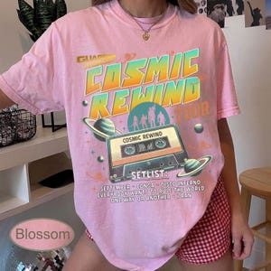 K&ouml;nnte beinhalten: Ein rosa T-Shirt mit einem Retro-Grafikdesign. Das Design zeigt die Worte "COSMIC REWIND TOUR" in Orange und T&uuml;rkis, eine Kassette, Planeten und eine Setlist mit Songs. Das T-Shirt ist mit "Blossom" beschriftet.