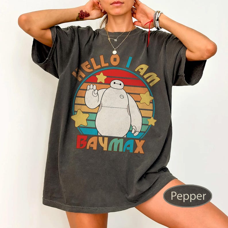 May include: Dark gray t-shirt featuring a graphic of Baymax from Big Hero 6. The design includes the text "HELLO I AM BAYMAX" in a circular design with stars and a retro color scheme. The shirt is a casual, oversized style.