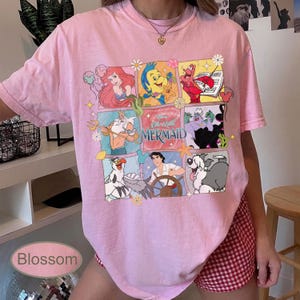 May include: Pale pink t-shirt with a colourful graphic of characters from Disney's "The Little Mermaid." The design includes Ariel, Flounder, Sebastian, and other characters in a grid pattern. The text "The Little Mermaid" is also visible.