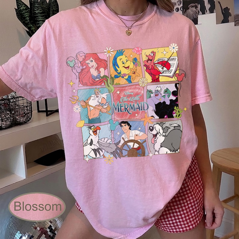 May include: Pale pink t-shirt with a colourful graphic of characters from Disney's "The Little Mermaid." The design includes Ariel, Flounder, Sebastian, and other characters in a grid pattern. The text "The Little Mermaid" is also visible.