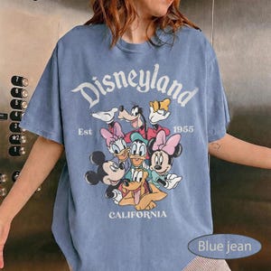May include: A blue jean-colored t-shirt featuring the word "Disneyland" in a white, vintage font. The shirt showcases a graphic of Disney characters including Mickey Mouse, Minnie Mouse, Donald Duck, and Goofy. The text "Est 1955" and "California" are also visible.