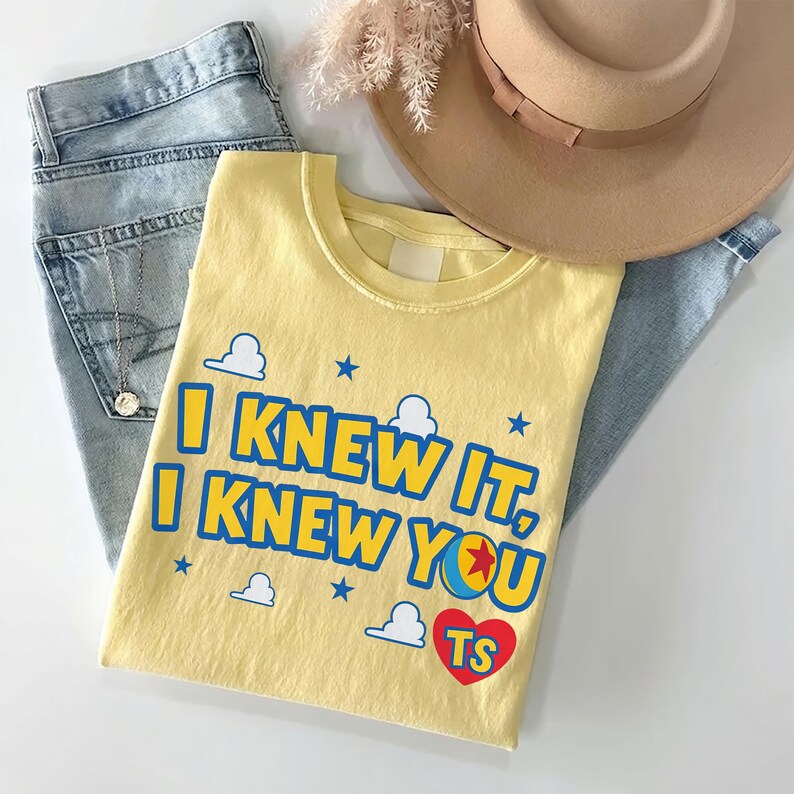 May include: A pale yellow t-shirt featuring the blue text I KNEW IT, I KNEW YOU with a red heart containing the letters TS, blue stars, and white cloud graphics. The shirt is displayed with light blue denim jeans, a tan hat, and a silver necklace.